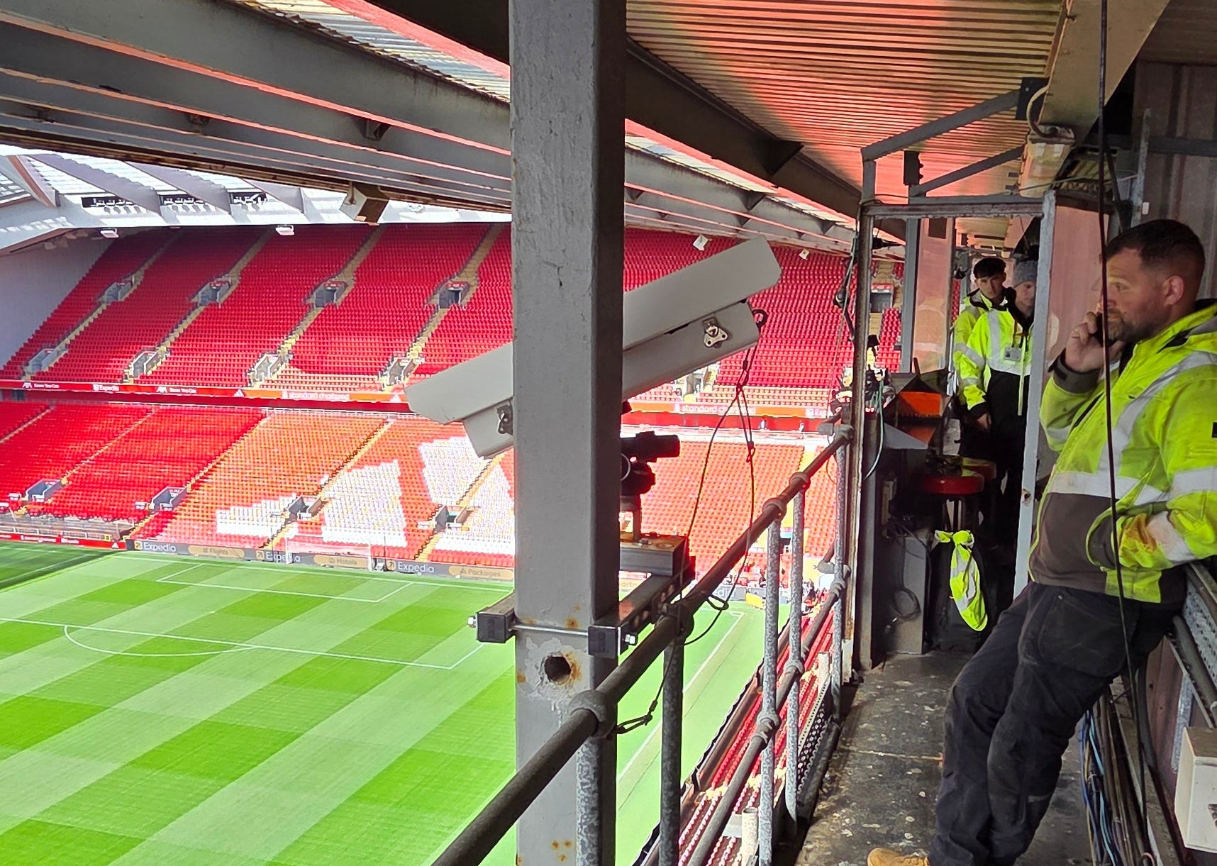 Stadium interior with green field and red seats, workers in high-visibility jackets.