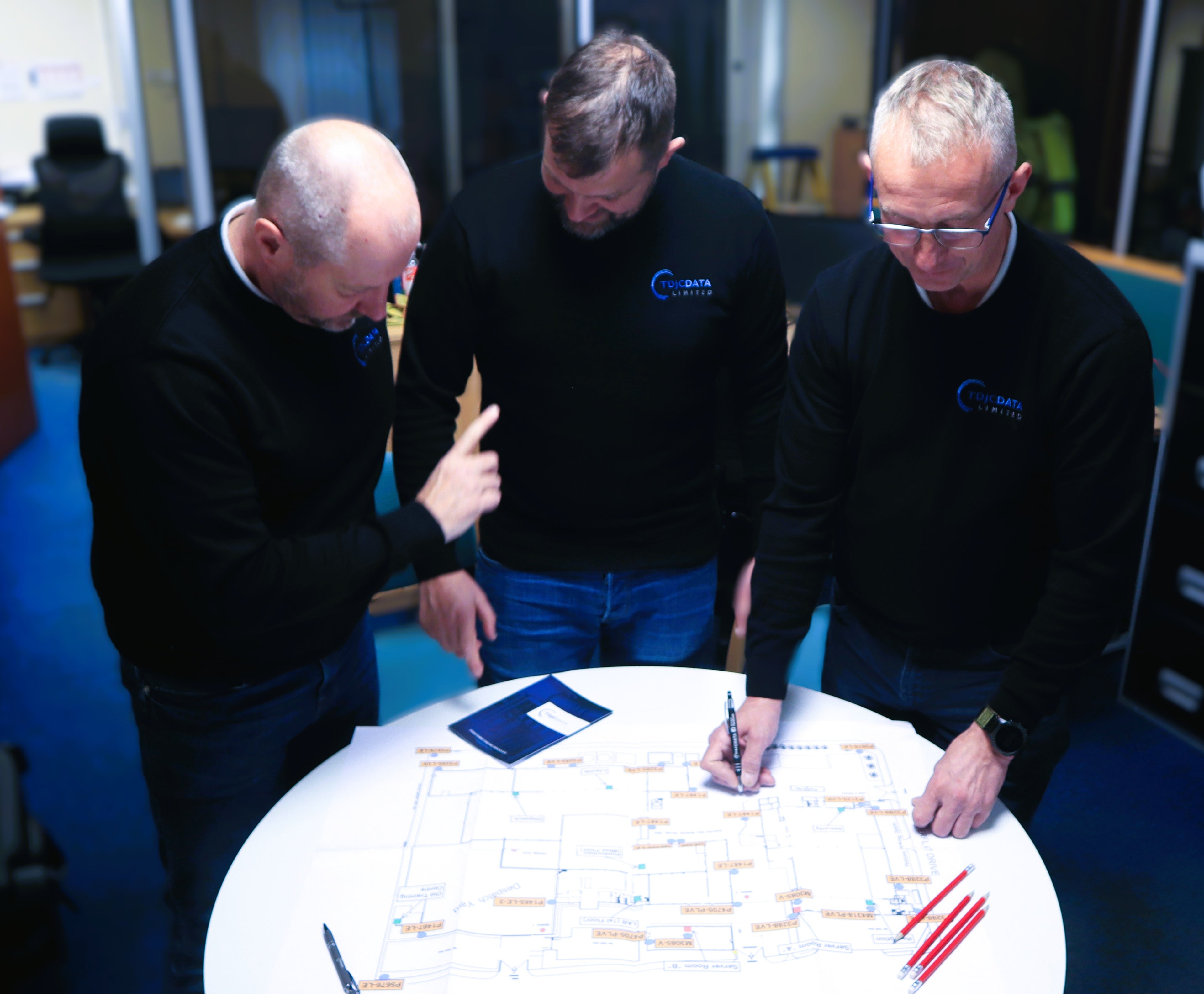 Three men in black jackets with logos discussing a document at a table.