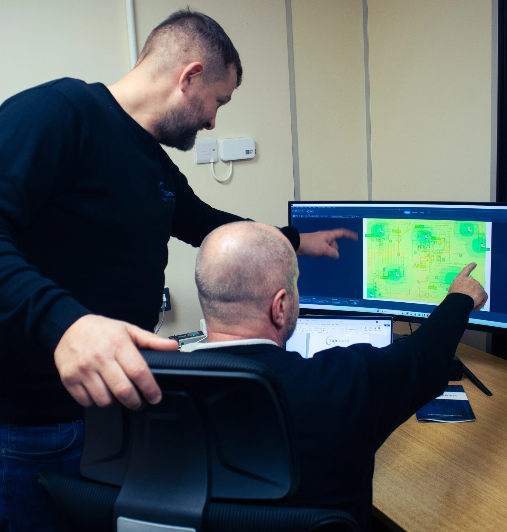 Two men working together at a desk with multiple computer monitors.